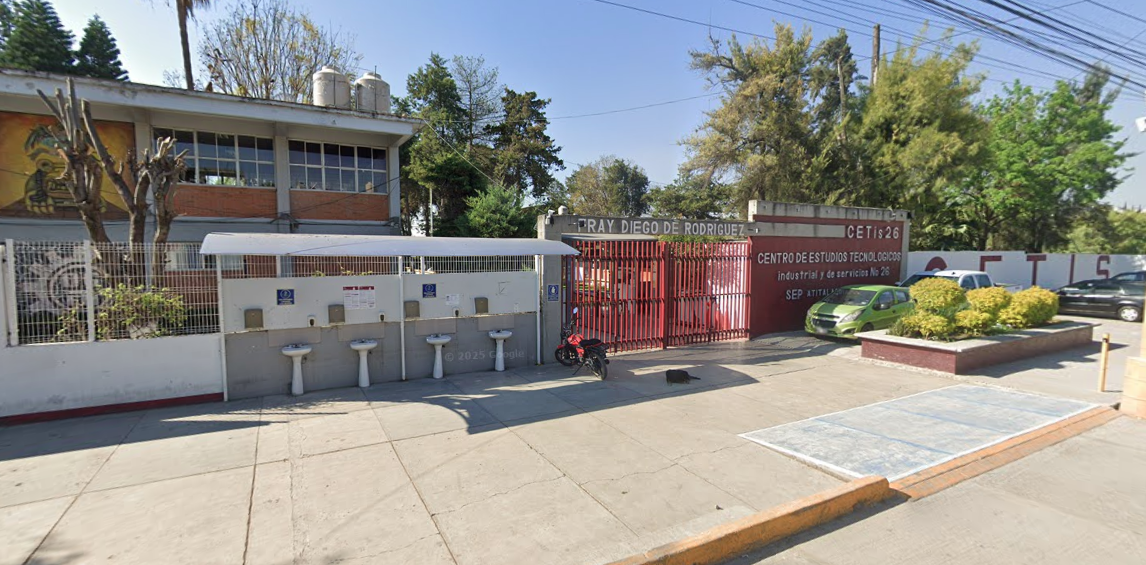 Fotografía en la que se muestra la entrada del plantel conformada por una reja de color rojo. A la izquierda de la reja hay un muro y frente a él hay cuatro lavabos blancos. Al lado derecha de la reja hay un muro rojo que en letras blancas dice CETIS 26.
